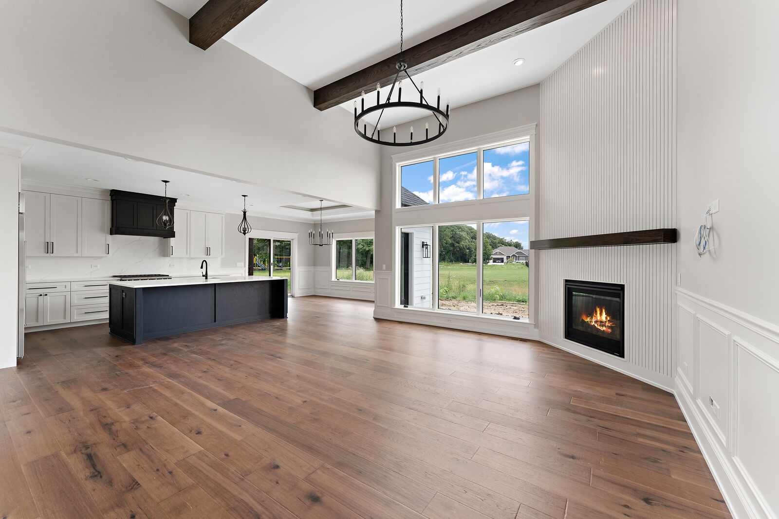 Vaulted great room with exposed wood beams, a wagon wheel chandelier and a fluted fireplace surround, open to the kitchen